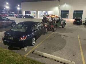 Black sedan and green tow truck at dealership night.