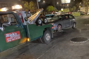 Nighttime green tow truck with gray hatchback.