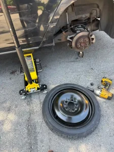 Spare wheel, floor jack and tools beside a vehicle with its wheel removed